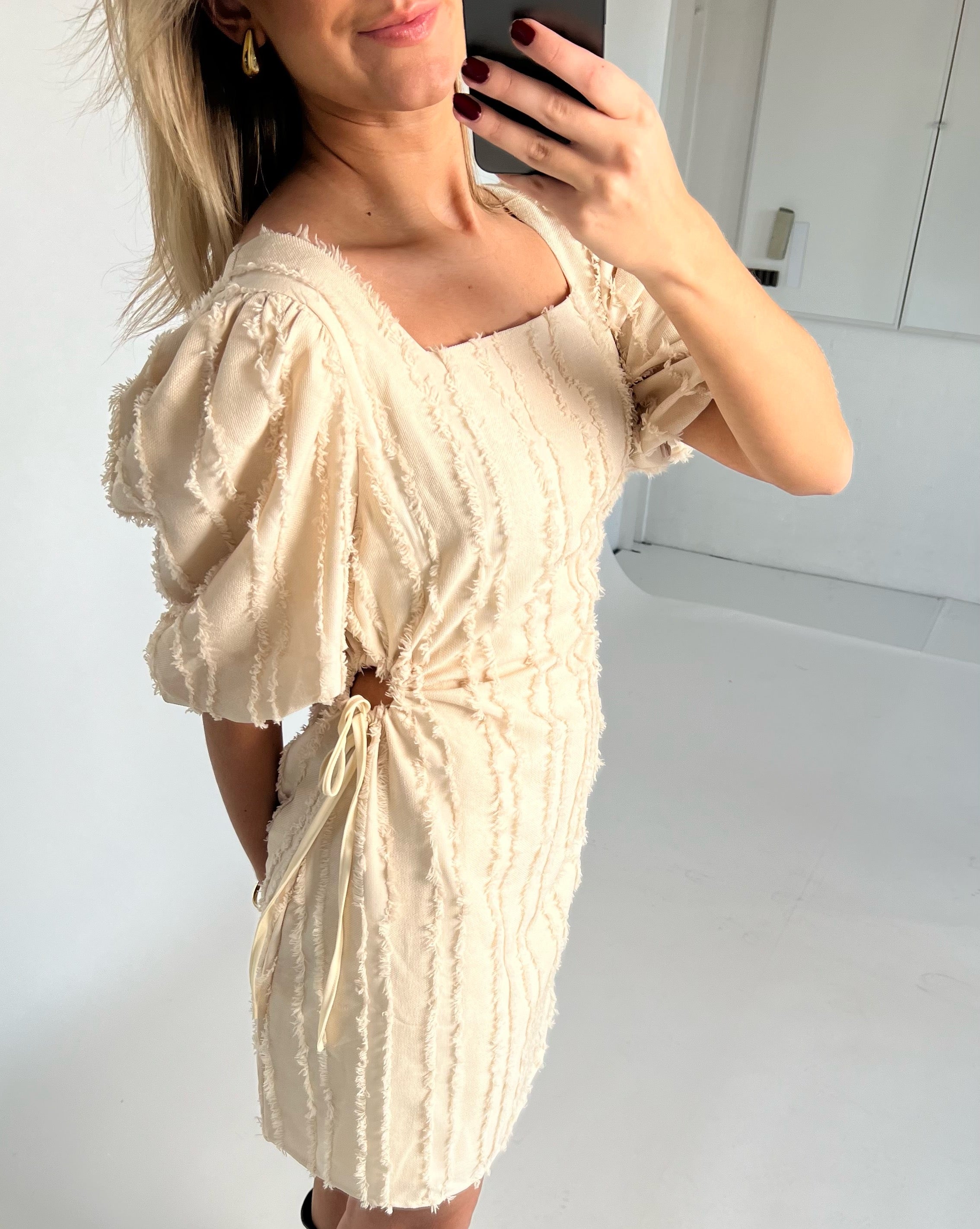 Kvinde iført off-white kjole med pufærmer tager et spejl-selfie. Spejlet er i et lyst, minimalistisk rum med en hvid væg og dør i baggrunden.