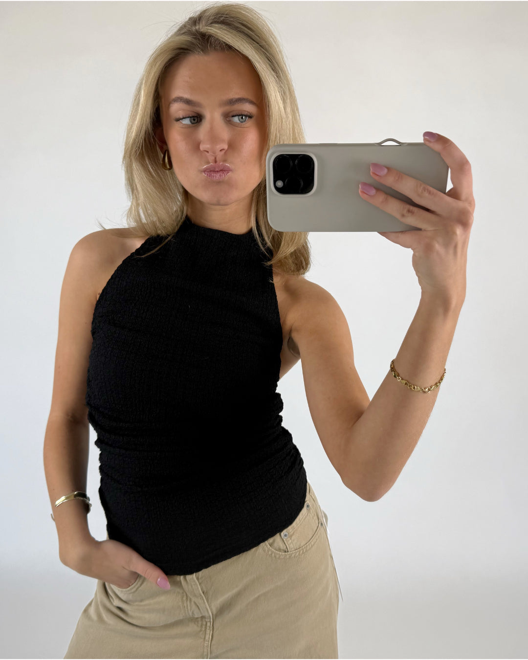 Kvinde tager en selfie med en smartphone, iført sort top og beige bukser. Hun poserer med et ansigtsudtryk og står i et lyst rum.