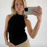 Kvinde tager en selfie med en smartphone, iført sort top og beige bukser. Hun poserer med et ansigtsudtryk og står i et lyst rum.