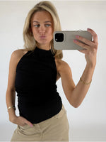 Kvinde tager en selfie med en smartphone, iført sort top og beige bukser. Hun poserer med et ansigtsudtryk og står i et lyst rum.