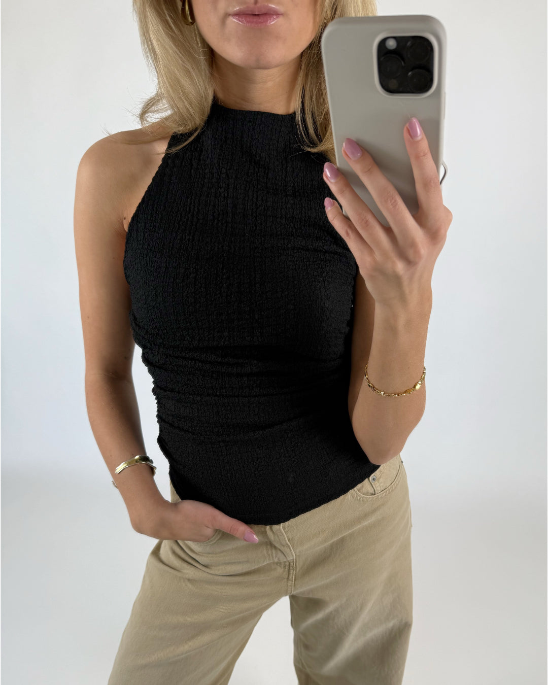 En person fotograferer sig selv med en smartphone iført en sort ærmeløs top og beige bukser. Personen står i et minimalistisk rum og bærer guldsmykker.
