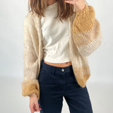 En person tager et spejlfoto. De har en beige og brun strikket cardigan over en hvid T-shirt og mørkeblå bukser. Baggrund er simpel og lys.