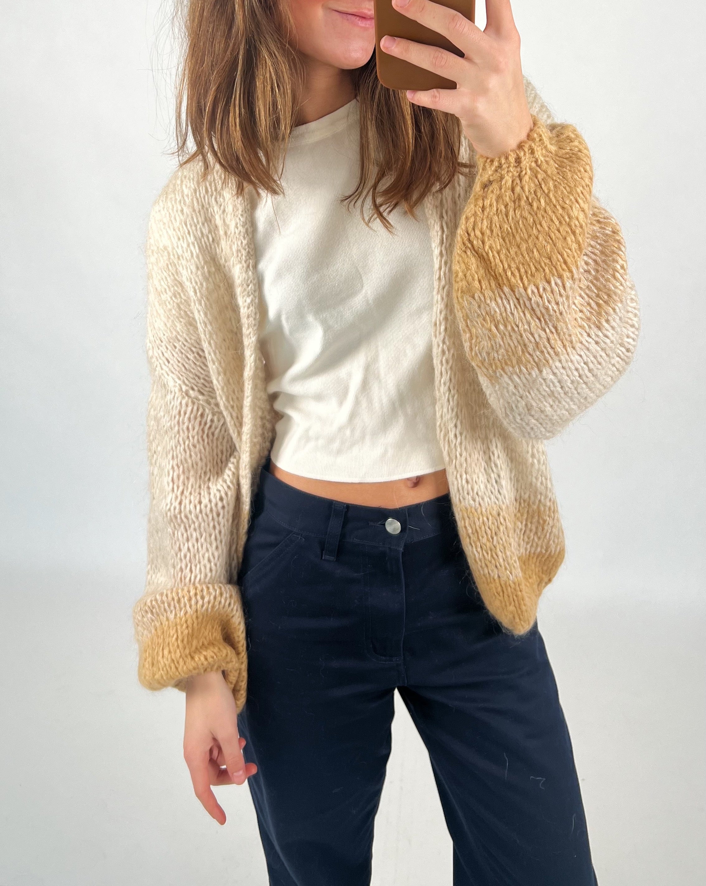 En person tager et spejlfoto. De har en beige og brun strikket cardigan over en hvid T-shirt og mørkeblå bukser. Baggrund er simpel og lys.