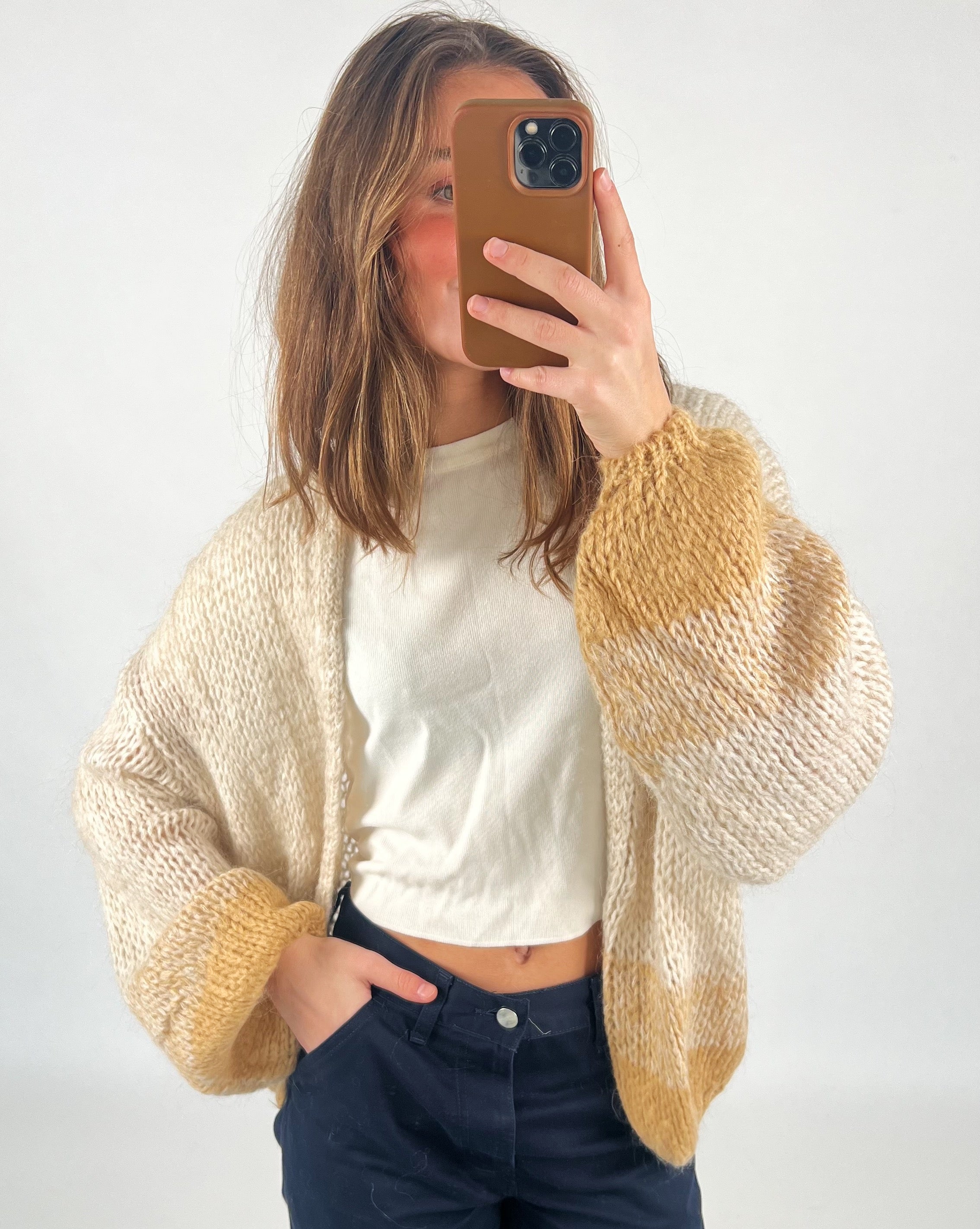 En person tager et spejlbillede iført en strikket cardigan med beige og guldfarvet mønster og en hvid t-shirt. Personen står i et enkelt, lyst rum.