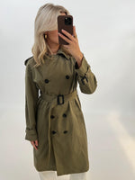 Person iført grøn trenchcoat tager et spejlbillede med en smartphone. Hun står i et minimalistisk rum med lyse vægge.