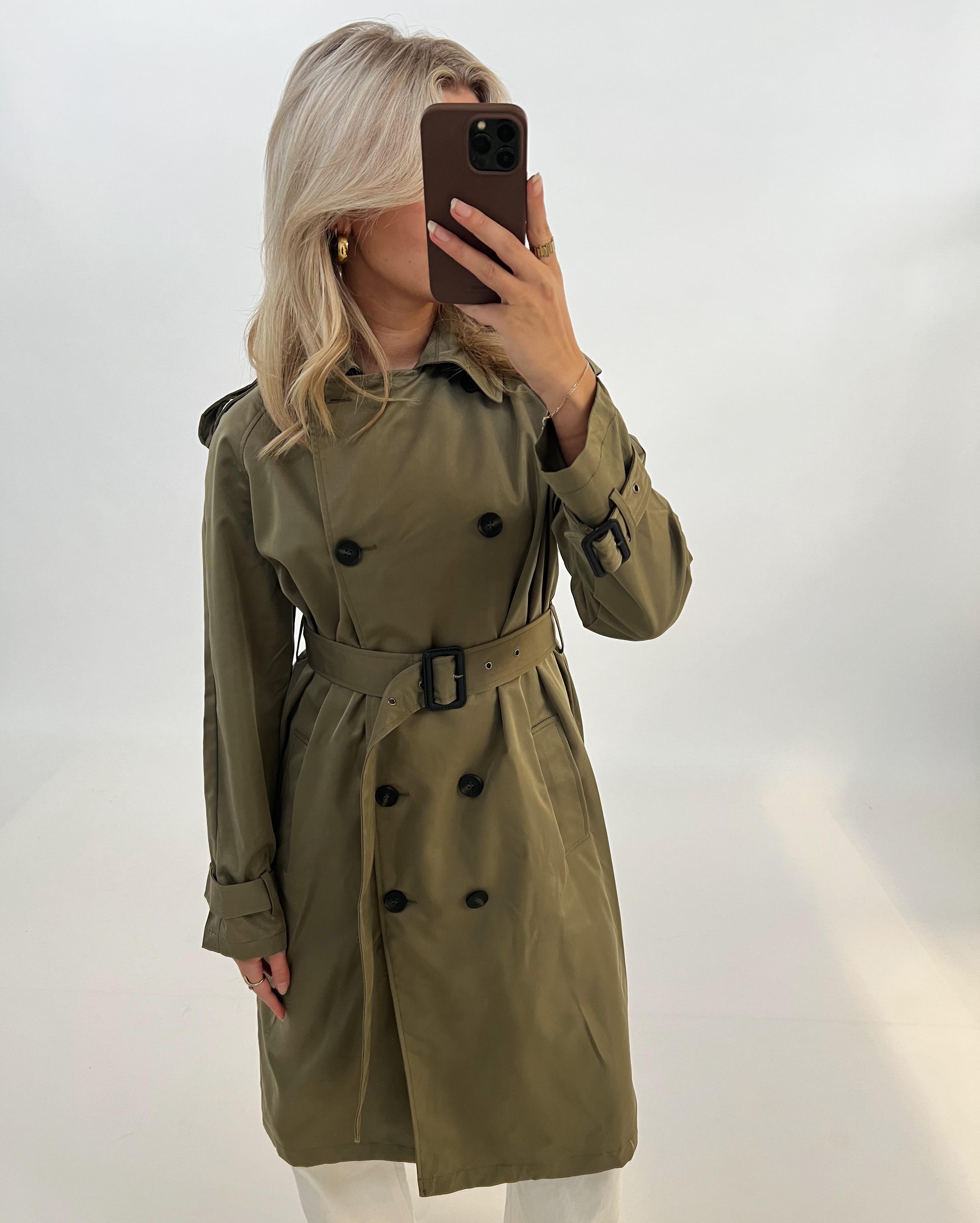 Person iført grøn trenchcoat tager et spejlbillede med en smartphone. Hun står i et minimalistisk rum med lyse vægge.