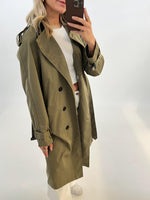 En person iført en grøn trenchcoat tager en selfie i et spejl. Personen bærer også en hvid trøje og bukser i en lys, minimalistisk baggrund.