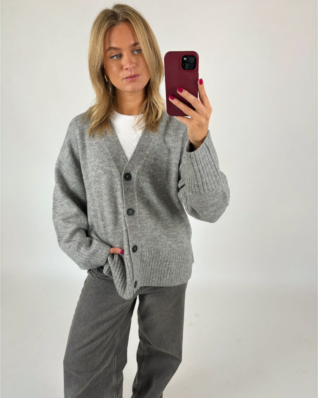 Person tager selfie, iført grå cardigan og mørke bukser; står i neutral, hvid baggrund. Personen har langt, lyst hår og holder en rød telefon med én hånd i lommen.