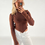 En person iført en brun sweater og hvide bukser tager et spejlbillede med en smartphone i en enkel, lys baggrund.
