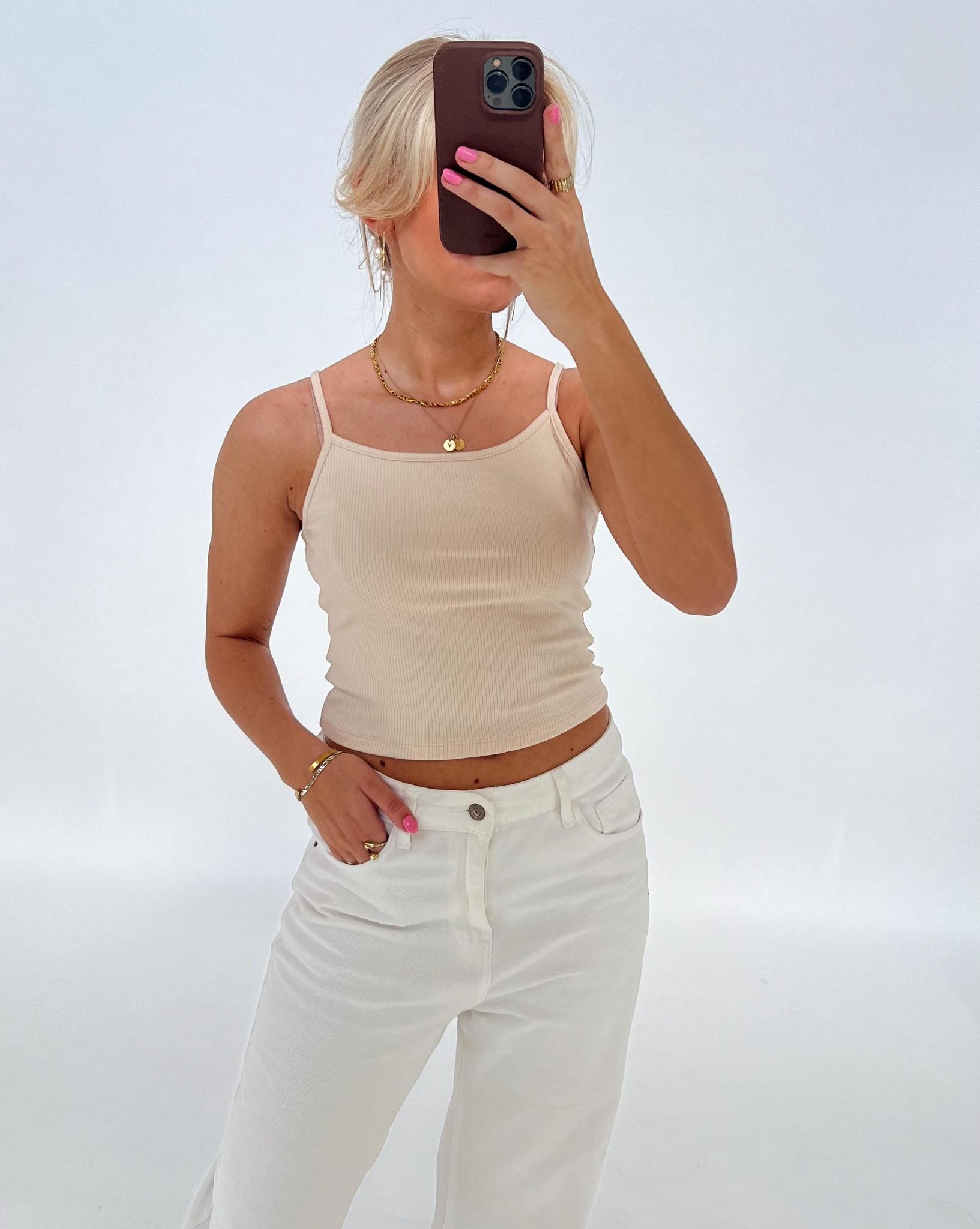 Person tager et selfie; iført beige top og hvide bukser i et lyst rum. Personen holder telefonen op foran ansigtet og har neglelak på fingrene.