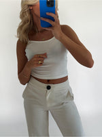 En person tager et spejl-selfie iført en hvid top og hvide bukser. Personen holder en blå telefon, og omgivelserne er minimalistiske med en hvid baggrund.