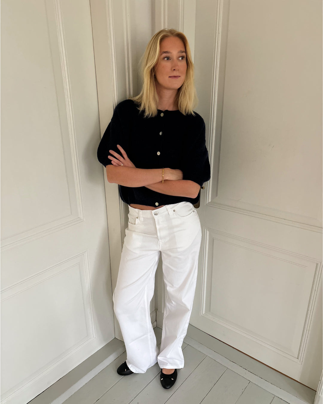 En person står afslappet med korslagte arme iført en sort trøje og hvide bukser. Baggrunden består af lyse paneldøre, hvilket giver et minimalistisk og stilfuldt look.