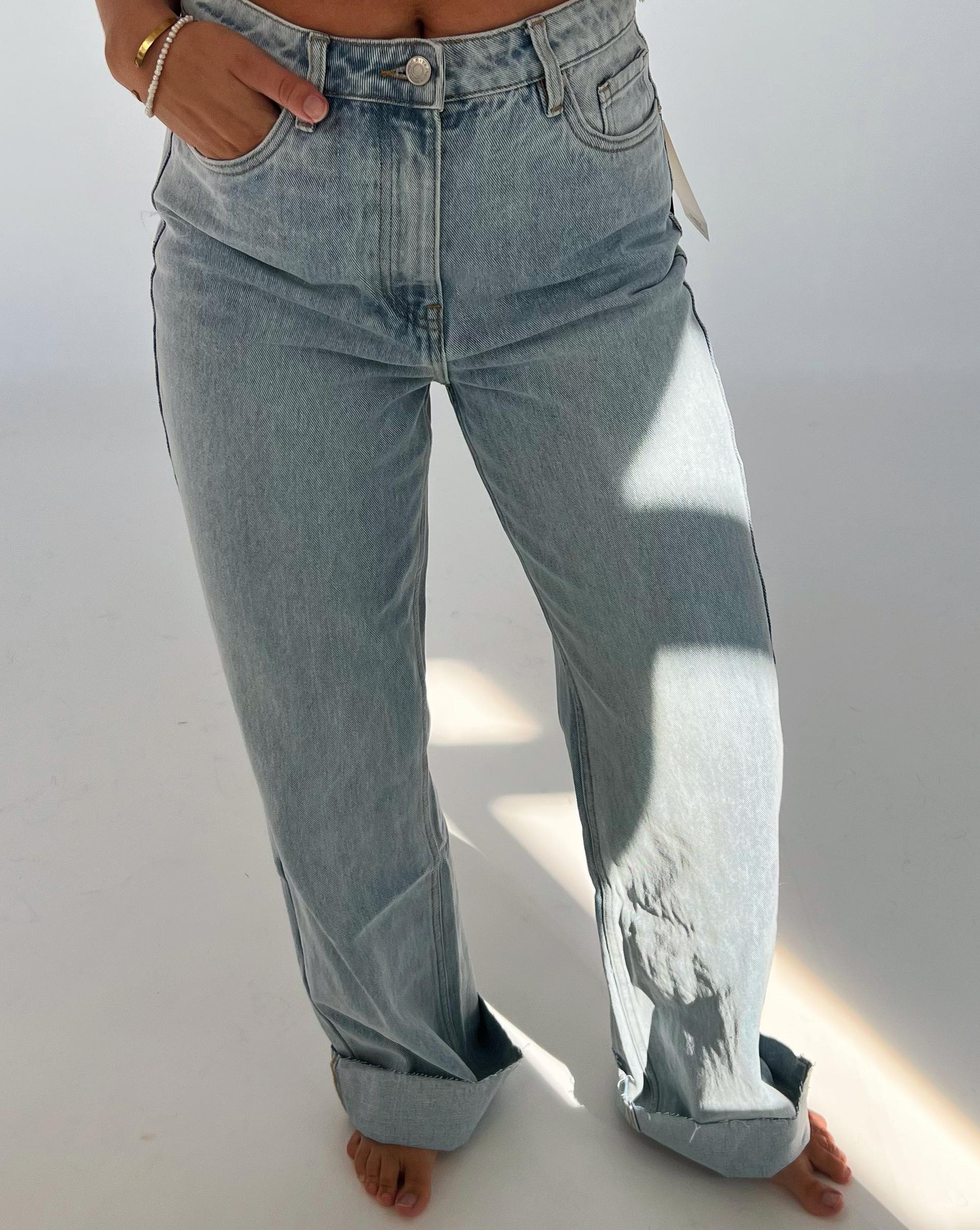 Person iført lyseblå jeans står med hænderne i lommerne i et lyst rum. Bunden af bukserne er rullet op, og der er solskin på den ene side.