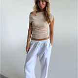 En person står afslappet iført en beige off-shoulder top og hvide bukser. Personen har hænderne i lommerne og befinder sig i et lyst, minimalistisk rum.