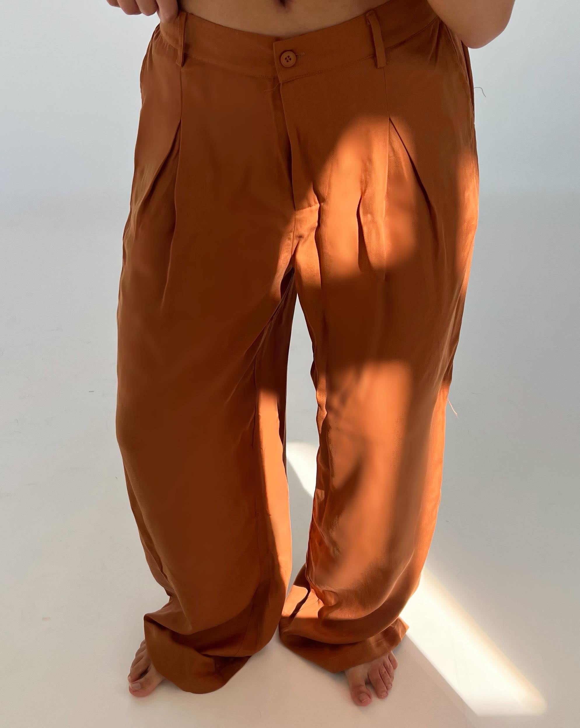 Brune bukser hænger løst på en person, der står barfodet. Sollys falder på stoffet, fremhæver folderne, i et lyst, minimalistisk rum.