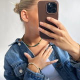 En person holder en telefon i en brun etui foran ansigtet, iført en denimjakke, grøn halskæde, og flere armbånd, i et neutralt rum.