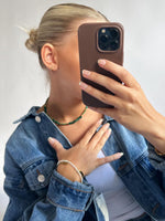 En person holder en telefon i en brun etui foran ansigtet, iført en denimjakke, grøn halskæde, og flere armbånd, i et neutralt rum.