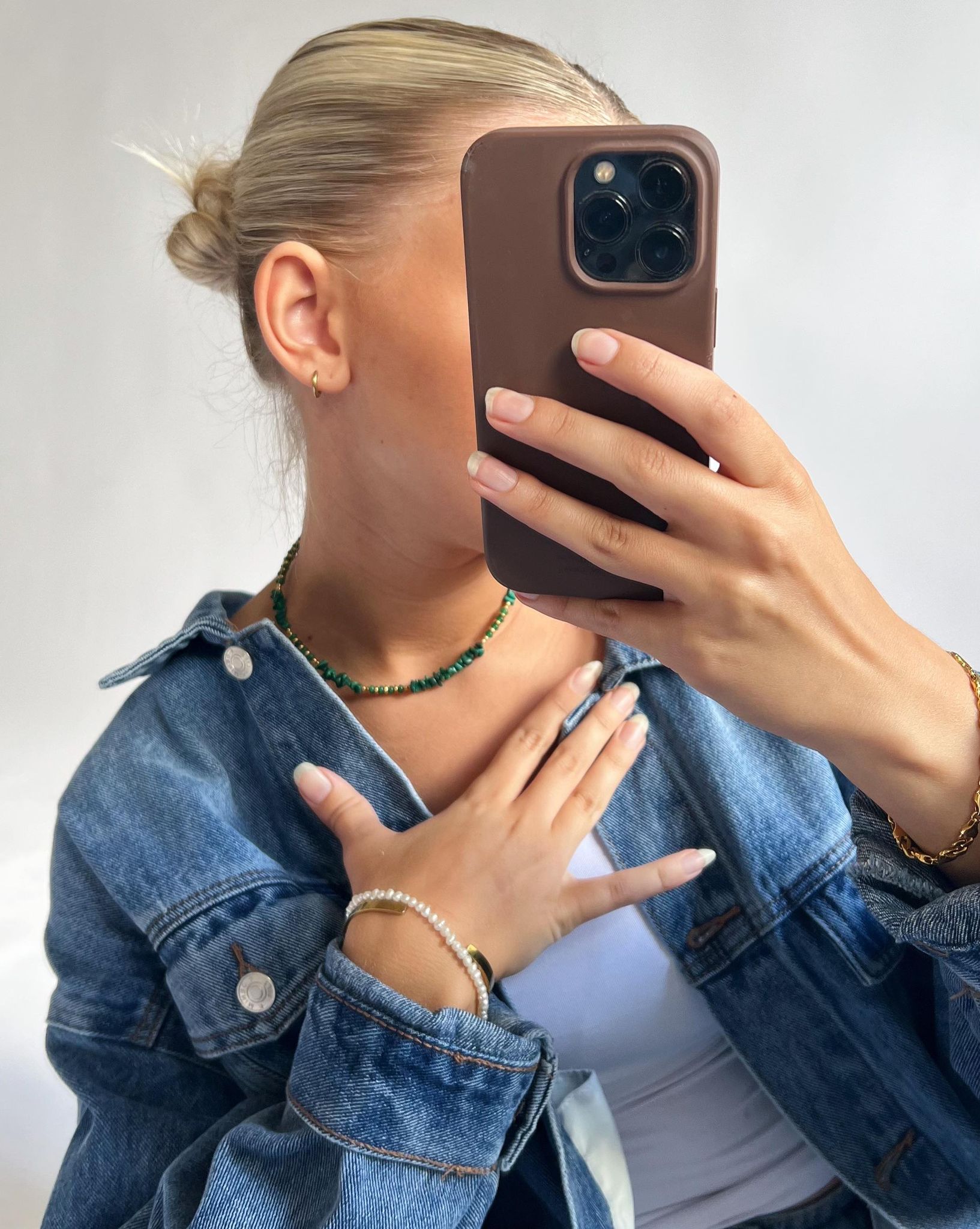 En person holder en telefon i en brun etui foran ansigtet, iført en denimjakke, grøn halskæde, og flere armbånd, i et neutralt rum.