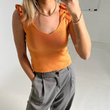 En person iført en orange top med flæser og grå bukser tager et selfie i en hvidmalet indendørs setting.
