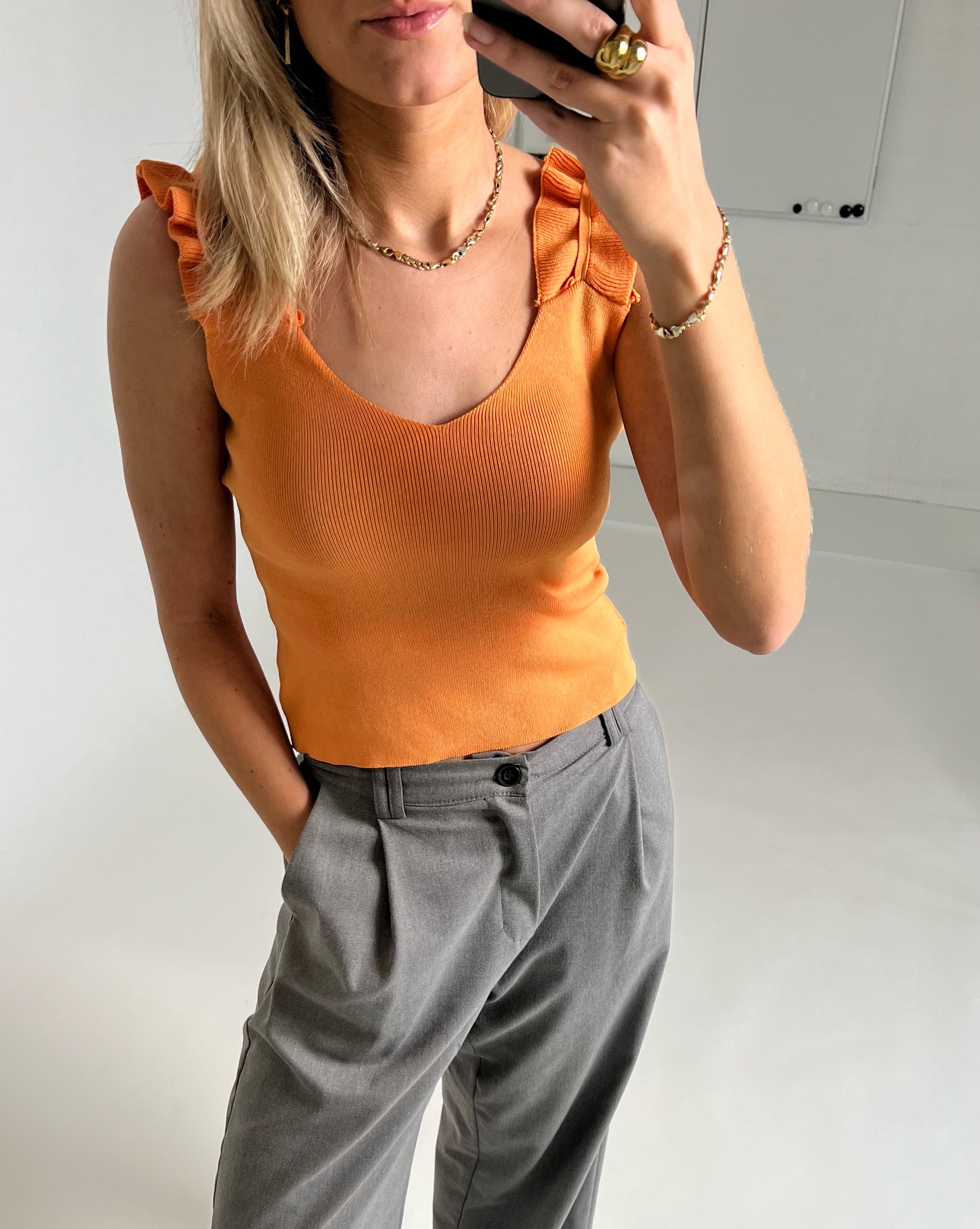 En person iført en orange top med flæser og grå bukser tager et selfie i en hvidmalet indendørs setting.