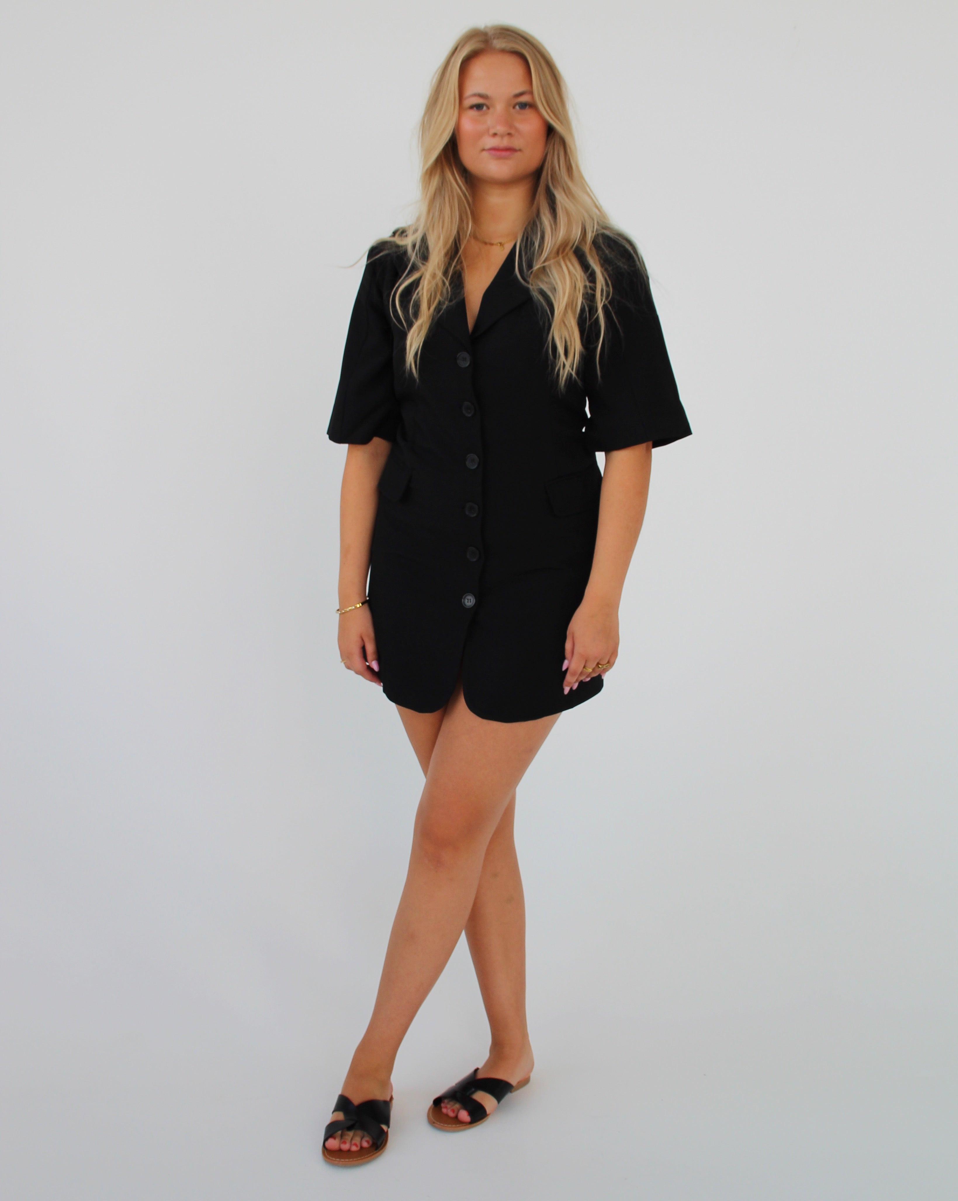 En person står med armene afslappet, iført en sort kortærmet kjole og sandaler. Baggrunden er en ensfarvet, lys væg, hvilket giver et minimalistisk look.