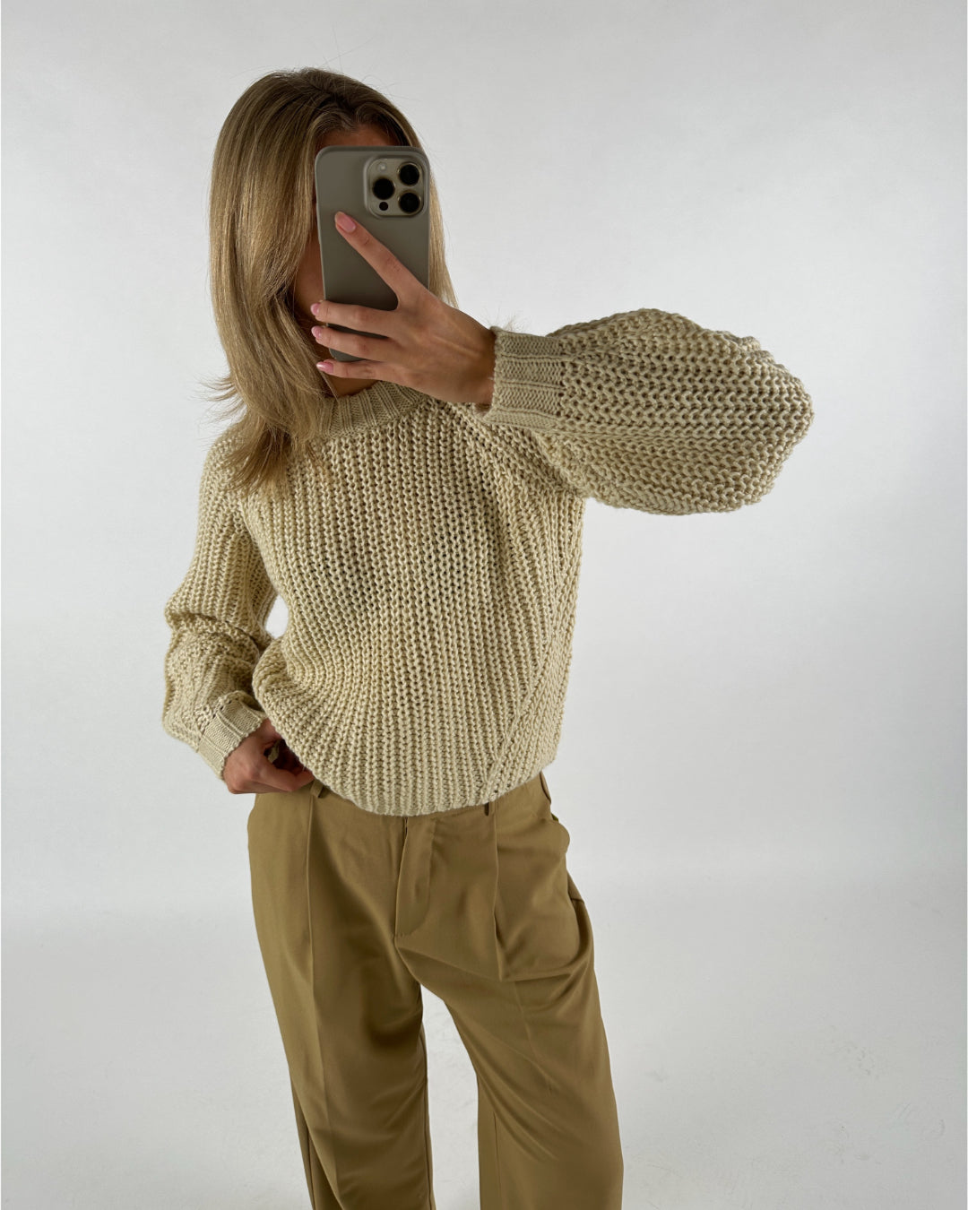 En person tager en selfie i spejlet iført en strikket, cremefarvet sweater med lange ærmer og beige bukser i et minimalistisk rum.