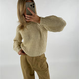 En person tager en selfie i spejlet iført en strikket, cremefarvet sweater med lange ærmer og beige bukser i et minimalistisk rum.