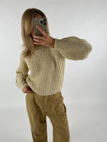 En person tager en selfie i spejlet iført en strikket, cremefarvet sweater med lange ærmer og beige bukser i et minimalistisk rum.
