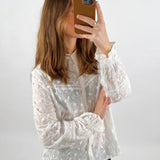 Person tager selfie med mobiltelefon iført hvid, broderet bluse. Baggrunden er enkel og lys, hvilket fremhæver det delikate mønster på blusen.