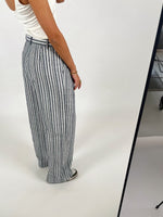 En person iført hvid tanktop og blå-hvid stribede bukser står mod en lys baggrund, mens de poserer i et fotostudie med hjul under en bordkant.