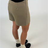 En person iført beige shorts står oprejst, iført sorte sko og hvide strømper, på en lys baggrund. Personen bærer også en sort trøje.