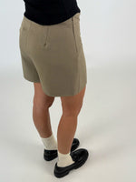 En person iført beige shorts står oprejst, iført sorte sko og hvide strømper, på en lys baggrund. Personen bærer også en sort trøje.