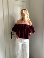 En person står i en gang iført en burgundy off-shoulder bluse og hvide bukser. Baggrunden viser en hvid dør og vægge.