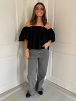 En person står afslappet iført en sort off-shoulder bluse og grå denim bukser i et rum med lyse vægge og gulve.