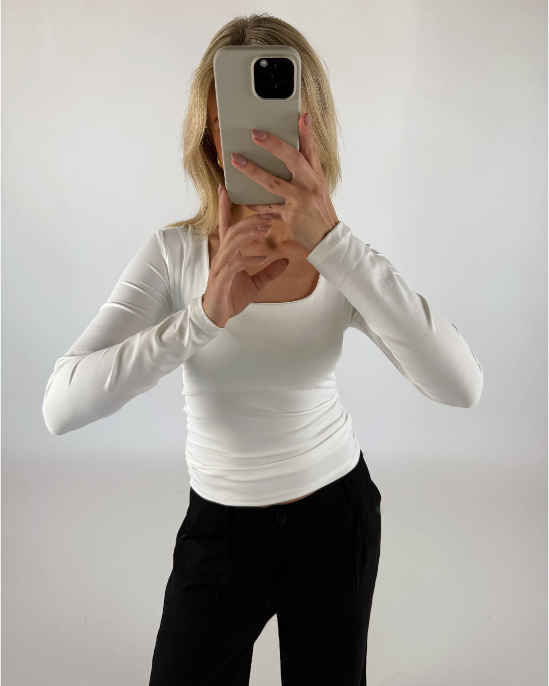 En person iført en hvid trøje holder en mobiltelefon op foran ansigtet i en lys, neutral baggrund.