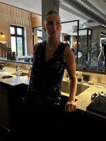 En person iført en glitrende sort top står ved en bar, omringet af glas og flasker, i en stilfuld café med dæmpet belysning og moderne indretning.