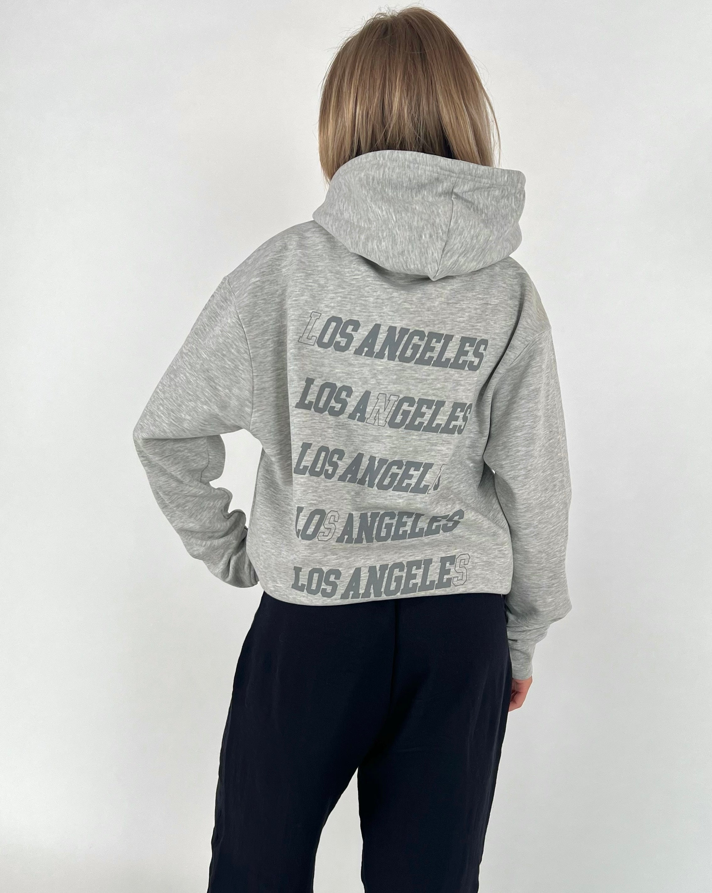 En person står med ryggen til og har en grå hættetrøje på, hvor der står "LOS ANGELES" fem gange. Personen er i et neutralt, lyst rum.