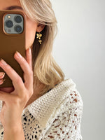 En person holder en mobiltelefon og ser i spejlet, iført en hvid strikket trøje og gyldne øreringe. Baggrunden er minimalistisk og lys.
