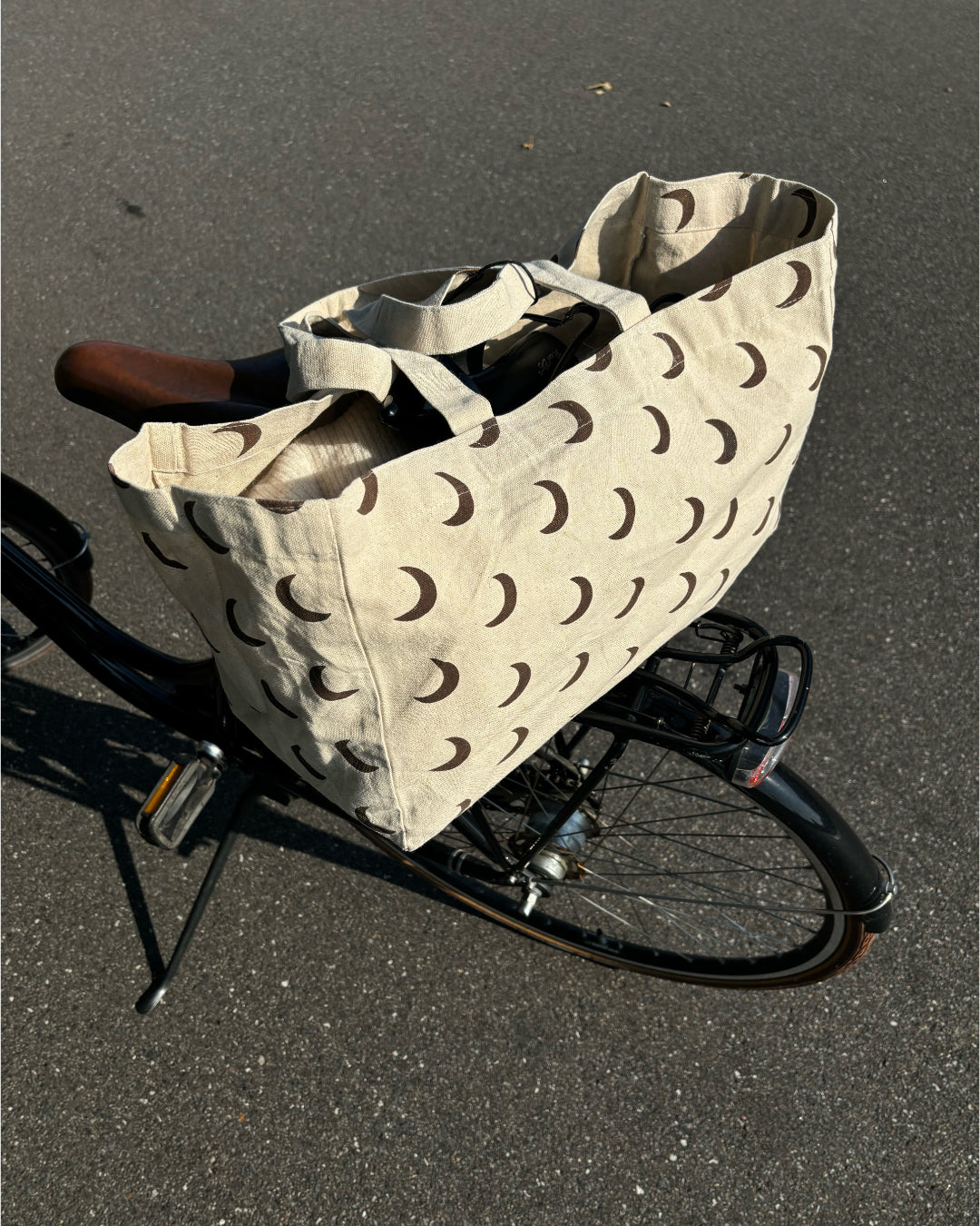 En beige stofpose med brune halvmåner, placeret på bagagebæreren af en cykel, står parkeret på en asfaltvej.