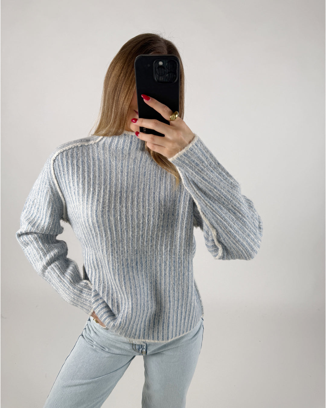 En person iført en lyseblå, stribet sweater tager et billede med en sort telefon. Personen er omgivet af en lys, neutralt farvet baggrund.