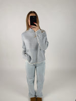 En person iført en lyseblå striktrøje og lyse jeans står i et minimalistisk rum, mens de tager et billede af sig selv med en smartphone.