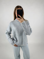 En person i en stribet sweater tager en selfie med en sort telefon. Personen står i et enkelt, hvidt rum og bærer afslappede, lyseblå jeans.