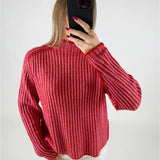 En person iført en rød- og pinkstribet sweater tager et spejlbillede med en sort mobiltelefon. Personen har rød neglelak og bærer en ring. Baggrunden er enkel og lys.