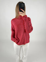 Person fotograferer sig selv, iført en rødstribet sweater og hvide bukser, med en mobiltelefon foran ansigtet. Baggrunden er enkel og lys.