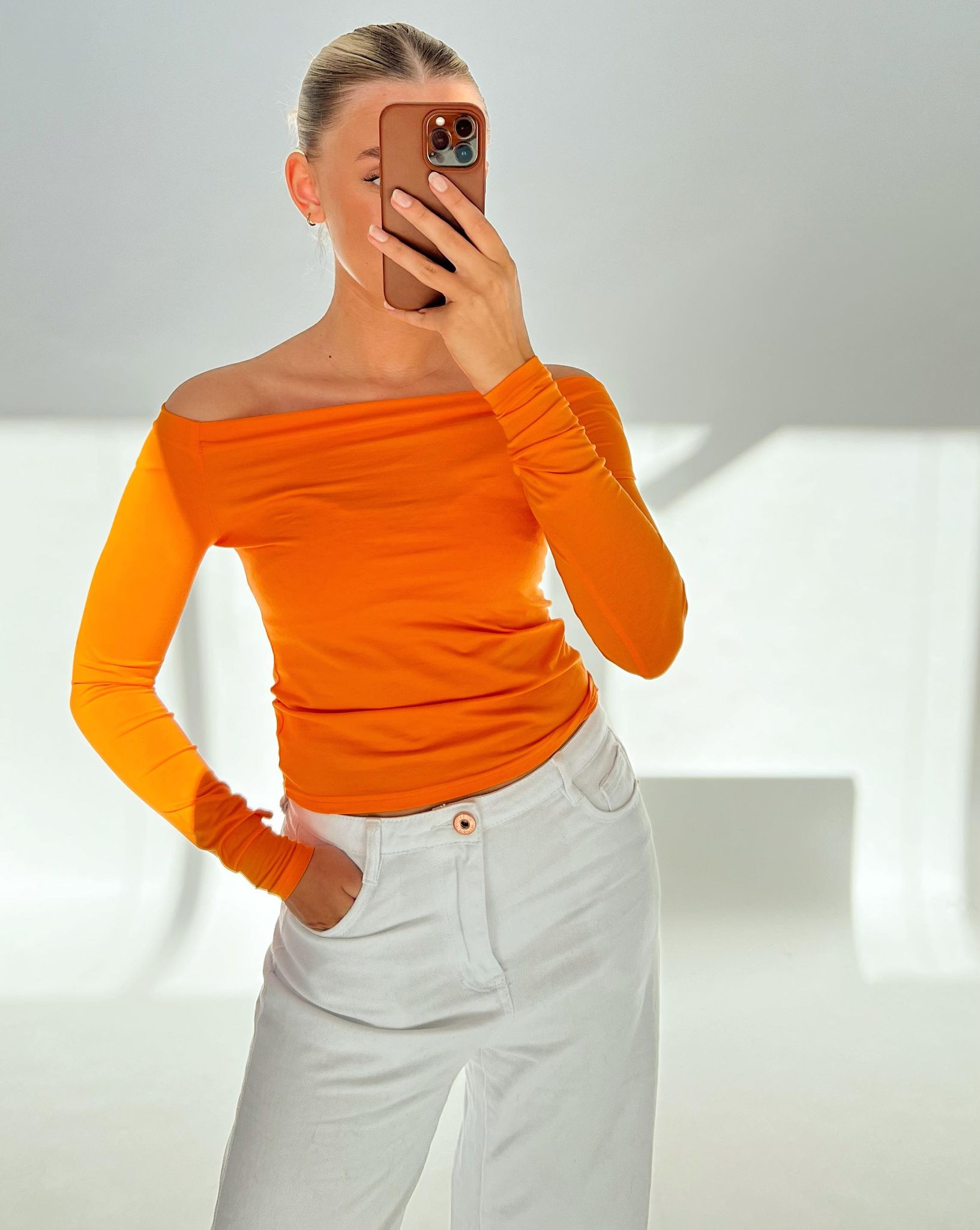 En person iført en orange off-shoulder trøje og hvide bukser tager en selfie i et lyst, hvidt rum. Personen har en telefon i en brun etui.