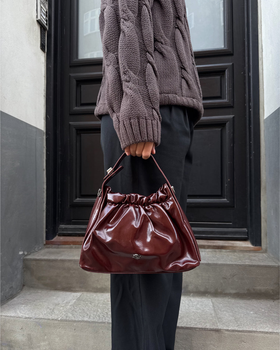 Modelfoto af pige med bordeaux Mallory taske i hånden. Hun er iført sorte bukser og brun Bay striktrøje. Alt er fra BySofieSønderby