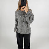 En person iført en grå, strikket sweater holder en sort telefon foran ansigtet. Personen står i et neutralt rum med en hvid baggrund.