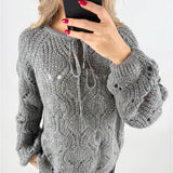 En person iført en grå, strikket sweater tager en selfie med en sort smartphone. Sweateren har snørebånd og et åbent mønster. Personen har røde negle, og baggrunden er neutral.