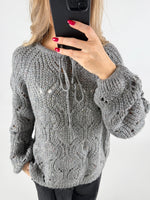En person iført en grå, strikket sweater tager en selfie med en sort smartphone. Sweateren har snørebånd og et åbent mønster. Personen har røde negle, og baggrunden er neutral.