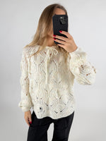 En person tager et spejlbillede iført en hvid kabelstrikket sweater og sorte bukser. Personen holder en sort smartphone, mens baggrunden er neutral og lys.